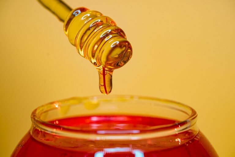 Golden honey dripping from a wooden dipper into a glass jar